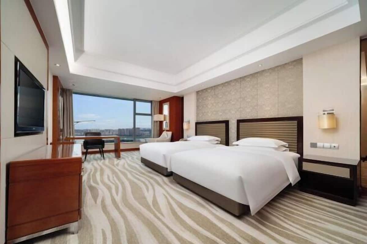 Habitación de hotel con dos camas, un escritorio, una silla, un televisor y un ventanal con vistas a la ciudad.