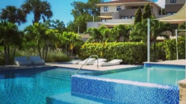 Una piscina con una fuente de agua cristalina azul, rodeada de sillas de descanso y un edificio de varios pisos al fondo.