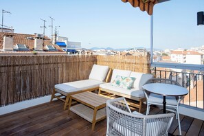 A balcony with a wooden floor, a sofa with cushions, a small table, and a wicker chair. - Studio Merlot - Welkeys (Cannes)