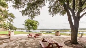 A stone patio with picnic tables and a large tree, overlooking a body of water.