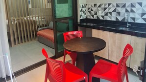 A small dining area with a round table and four red chairs, a sliding glass door leading to a balcony, and a kitchen area with a black countertop and a tiled backsplash. - Flat Térreo 02 Jardim by La Playa (Ipojuca)