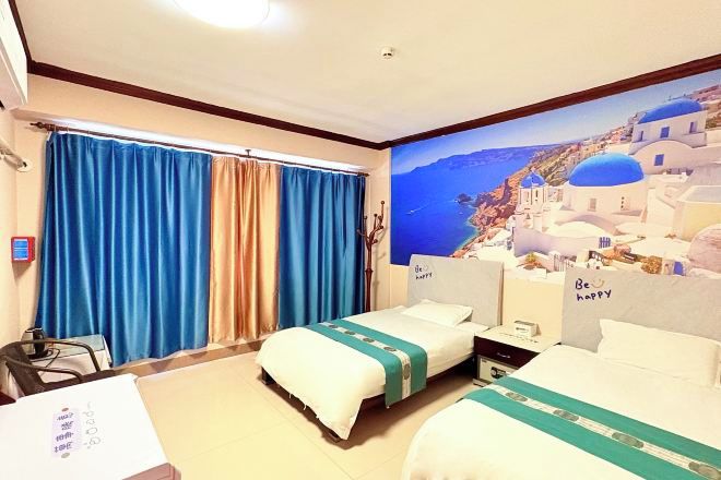 A hotel room with two beds, a large window with blue curtains, a painting of a coastal scene, and a sign on the headboard saying 'Be happy'.