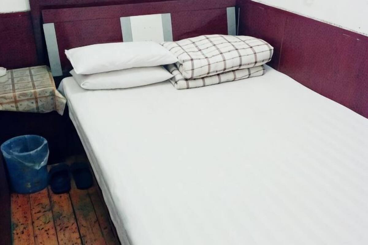 A single bed with white and plaid bedding, a blue trash bin, and a pair of black slippers on a wooden floor.