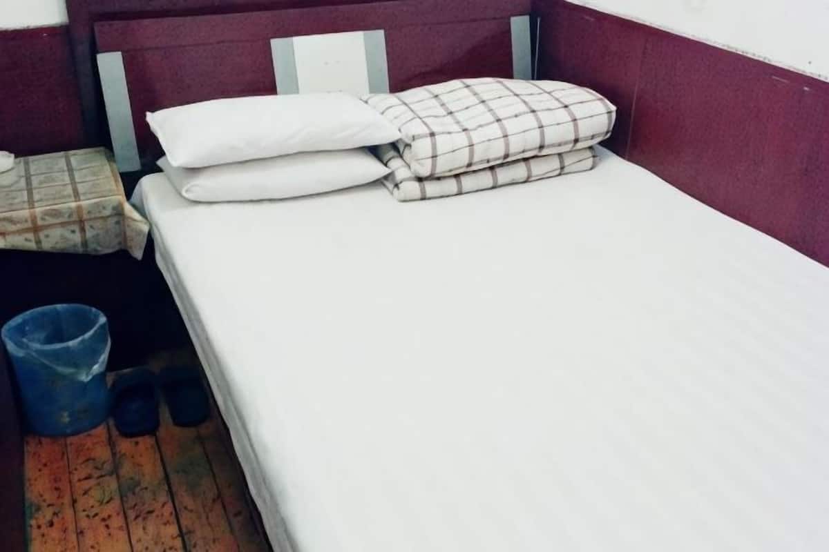 A single bed with white and plaid bedding, a blue trash bin, and a pair of black slippers on a wooden floor.