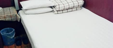 Un lit simple avec une literie blanche et à carreaux, une poubelle bleue et une paire de pantoufles noires sur un plancher en bois.