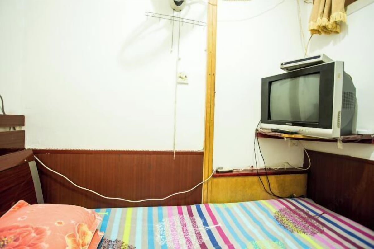A small, simple room with a bed, a television on a shelf, and a wooden headboard.