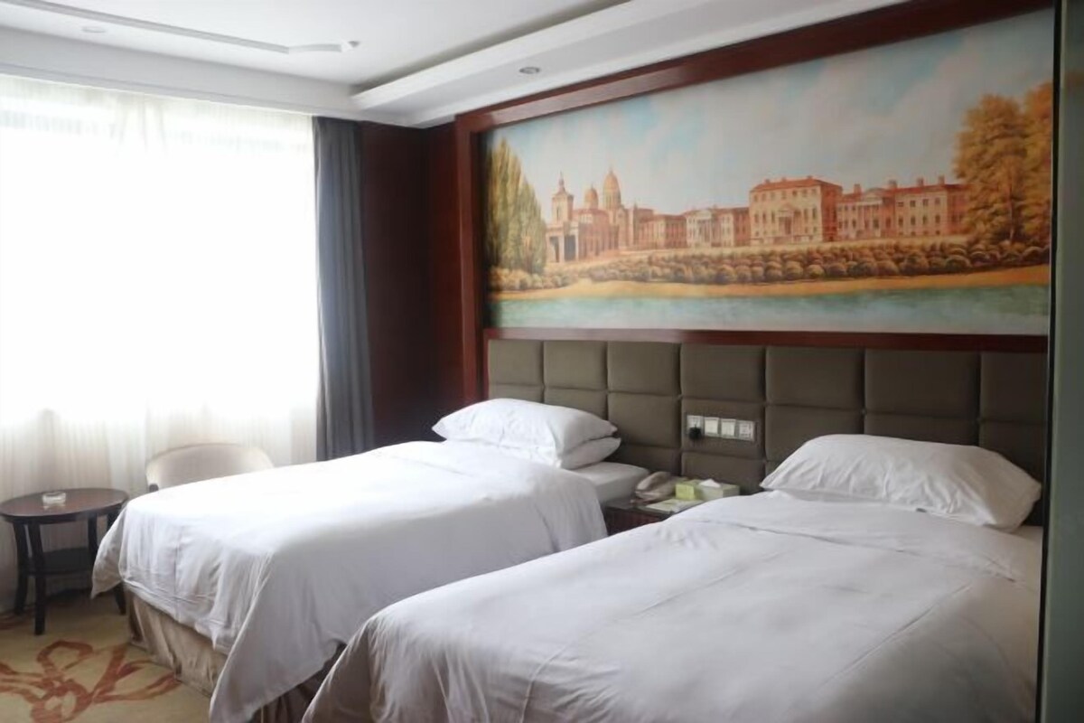 A hotel room with two beds, a painting of a cityscape, a small table, and a chair.