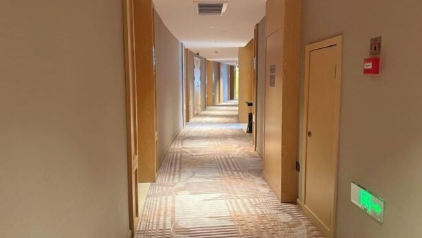 Un corridoio di un albergo con tappeto a motivi, porte in legno e un rilevatore di fumo sulla parete.