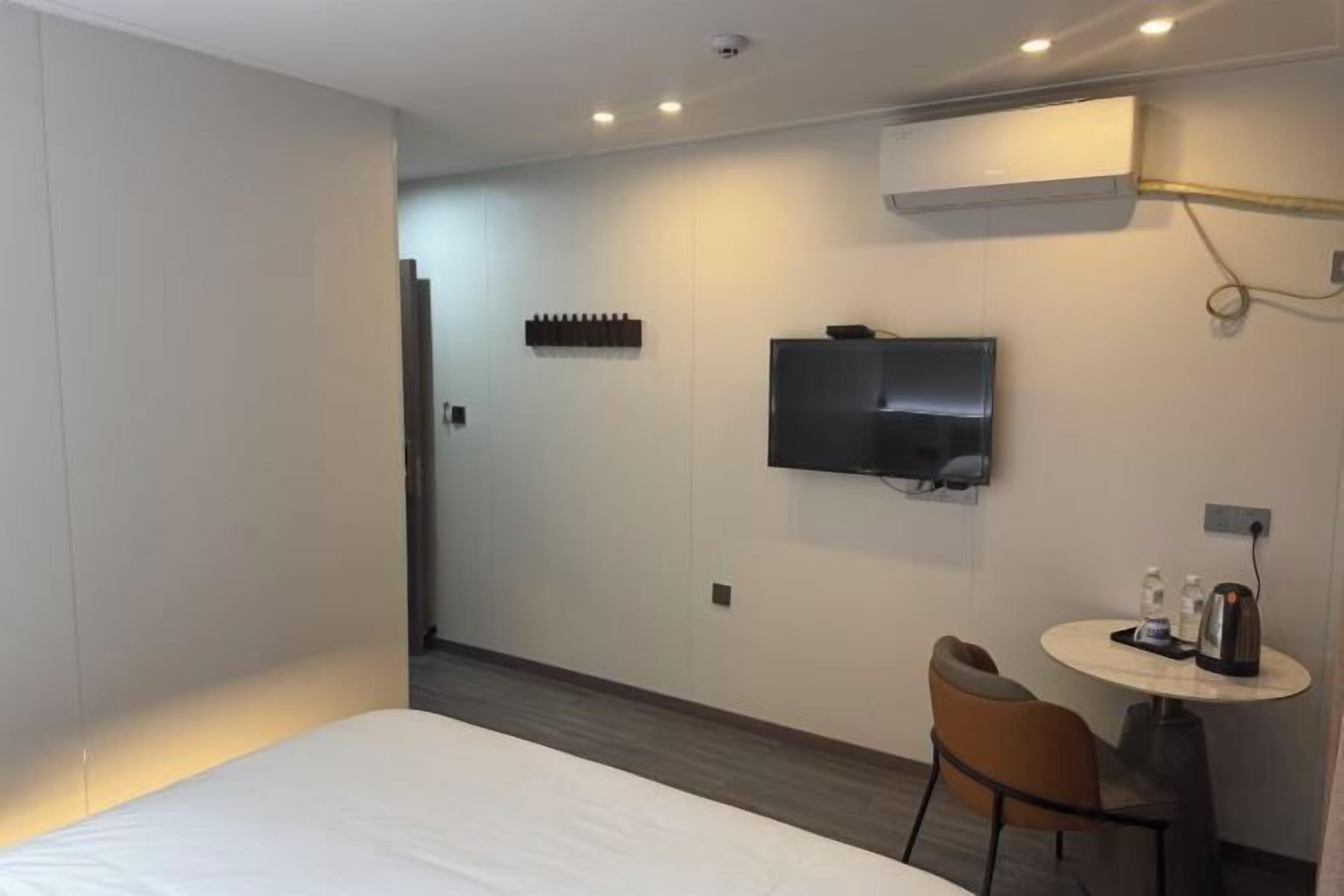 A hotel room with a bed, a TV mounted on the wall, a small table with a chair, and an air conditioning unit.