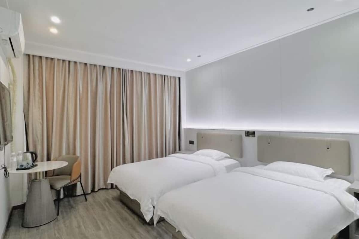 A hotel room with two beds, a small table with a chair, a wall-mounted air conditioner, and a curtain.
