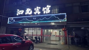 Entrada de un hotel con un letrero de neón, puertas de vidrio y un coche rojo estacionado afuera.