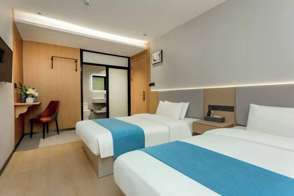 A hotel room with two beds, a desk, a chair, and a small bathroom.