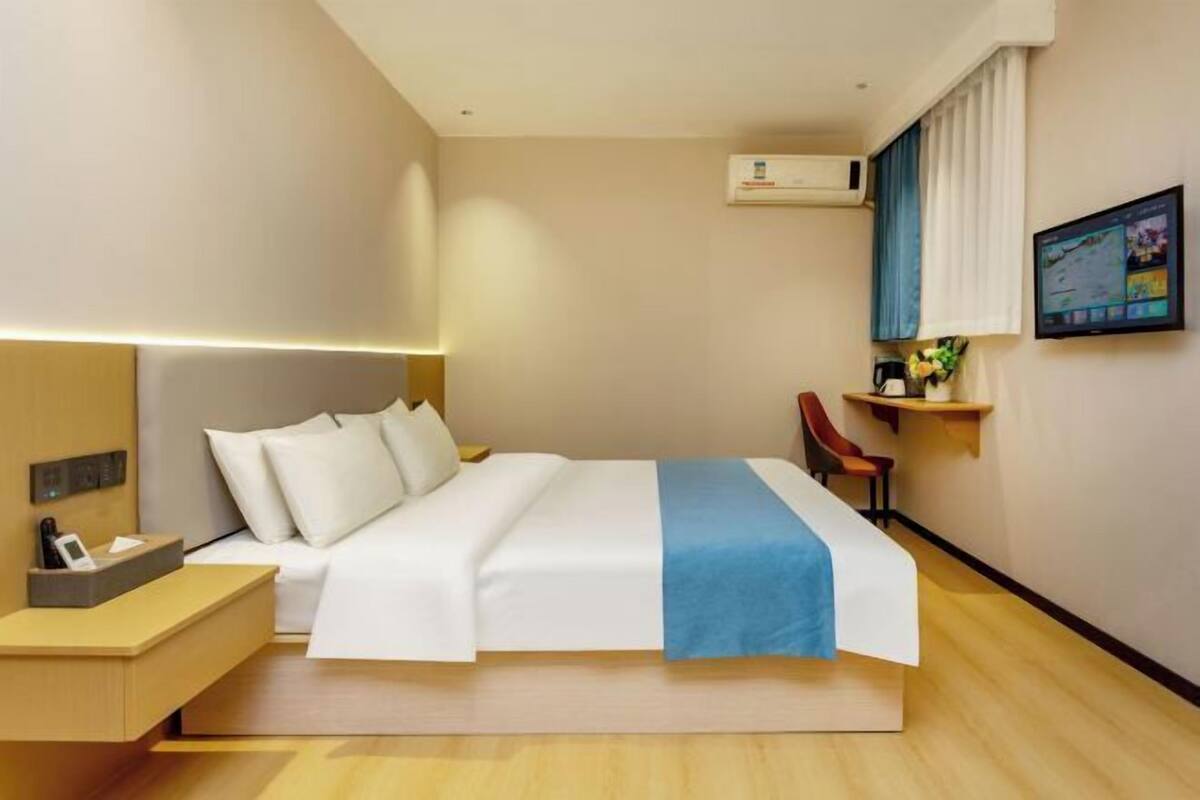 A hotel room with a bed, bedside table, desk, chair, and a TV on the wall.
