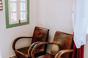 Una habitación con una silla de madera, un ventanal con detalles en verde y una cortina blanca.