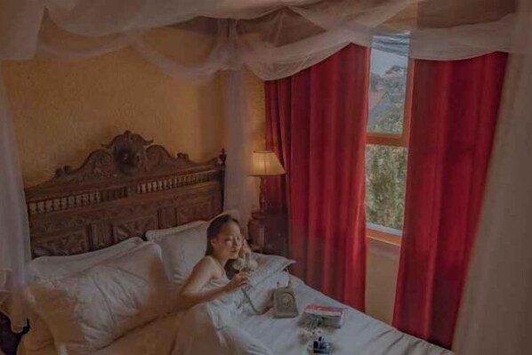 Un dormitorio con cama con dosel, una mesita de noche con lámpara y una ventana con cortinas rojas.