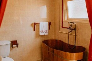 Un baño con una bañera de madera, un inodoro y una ventana con una toalla colgada en la pared.