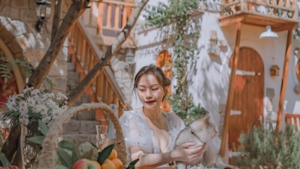 Une personne est assise, lisant un livre dans un cadre extérieur rustique, avec un mur de pierres, un balcon en bois et un panier de fruits.