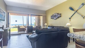 A living room with a black leather sofa, a dining area with a table and chairs, a view of the ocean through a sliding glass door, and a wall with a welcome sign and a life preserver.
