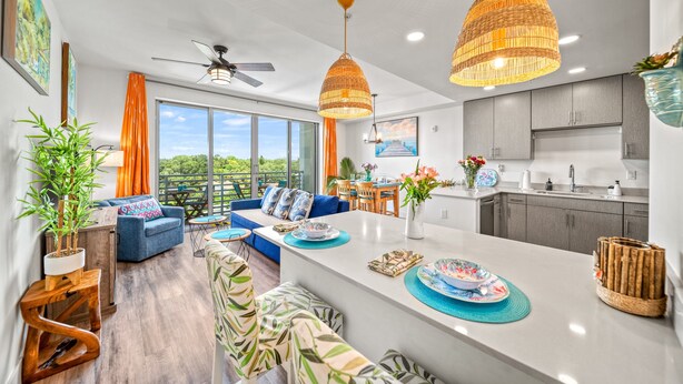 A modern kitchen with a central island, grey cabinets, and a dining area with a blue sofa and orange curtains. - Resort Pool & Spa, Fitness Center, Pet Friendly, Waterfront Condo in Downtown! (Sarasota)