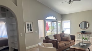 Ein Wohnzimmer mit braunem Ledersofa, einem weißen Sessel, einem Couchtisch und einem großen Spiegel. Ein Fenster mit Jalousien und eine Tür, die nach draußen führt.