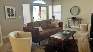 Ein Wohnzimmer mit braunem Sofa, einem weißen Sessel, einem Couchtisch und einem Teppich. An der Wand hängt ein Spiegel und auf dem Fensterbrett steht eine Pflanze.