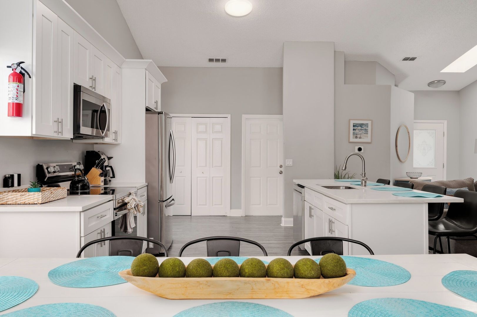 A modern kitchen with white cabinetry, a stainless steel refrigerator, and a microwave. There is a dining area with a table set for four, and a kitchen island with a sink and a bowl of green apples.