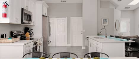 A modern kitchen with white cabinetry, a stainless steel refrigerator, and a microwave. There is a dining area with a table set for four, and a kitchen island with a sink and a bowl of green apples.