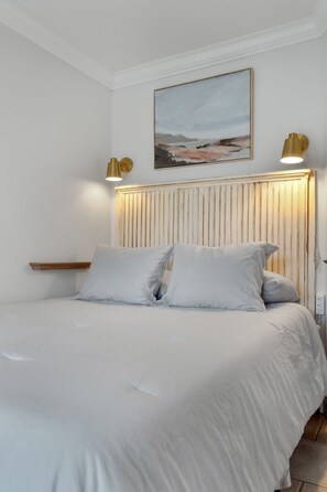 A bedroom with a bed, white bedding, a wooden headboard, and two wall-mounted lamps.