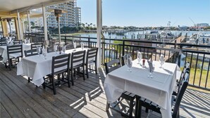 Un área de comedor al aire libre con mesas y sillas, con vistas a un puerto deportivo con barcos y edificios al fondo.