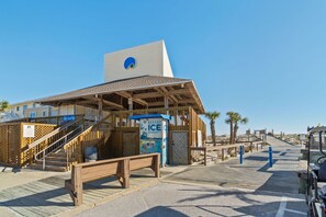 Ein Gebäude direkt am Strand mit einer Holzterrasse, einer Treppe zu einem überdachten Bereich und einem Automaten mit der Aufschrift 'BAGGED ICE'.