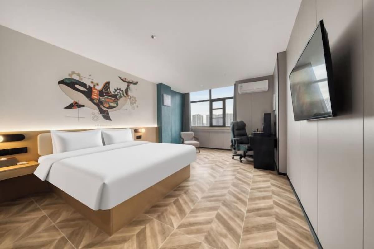 A modern hotel room with a large bed, a desk, a chair, a flat-screen TV, and a city view.