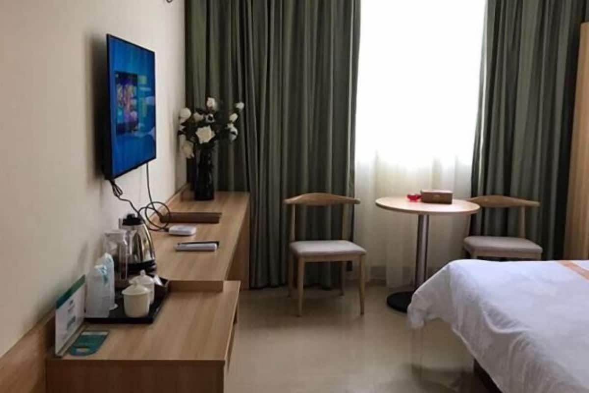 Una habitación de hotel con cama, televisor, una mesita con sillas, un jarrón con flores y una ventana con cortinas.