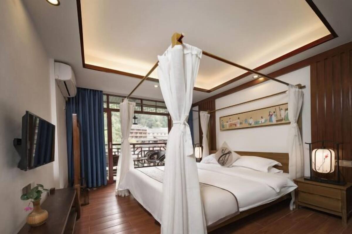 Une chambre à coucher avec un lit à baldaquin, une table de chevet, une commode, un téléviseur fixé au mur et une fenêtre avec des rideaux.