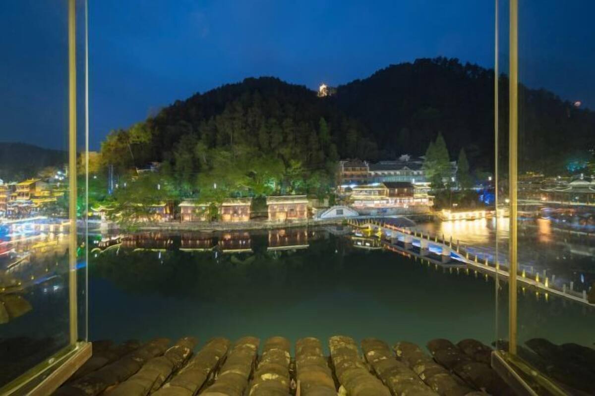 Une vue nocturne depuis une fenêtre donnant sur une rivière paisible, avec des bâtiments sur la rive opposée, un pont et une colline en arrière-plan.