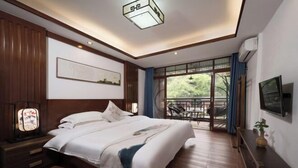 Ein Hotelzimmer mit einem großen Bett, Holzboden, einer Deckenleuchte, einem an der Wand montierten Fernseher und einem Fenster mit Blick auf Grünflächen.
