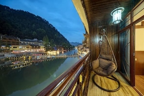A balcony with a hanging chair overlooking a river, wooden flooring, and a view of a town. - Baihuayuan River View Guesthouse (Fenghuang County)