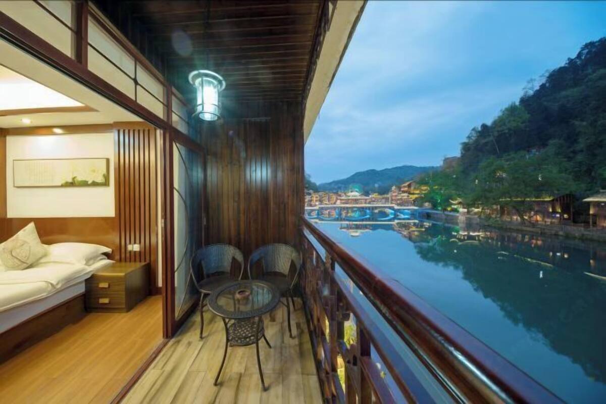 Un balcon avec un plancher en bois, un lit, une petite table et des chaises. On aperçoit une rivière et un pont au loin.