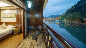 Ein Balkon mit Holzboden, einer Couch, zwei Stühlen und einem Geländer mit Blick auf einen Fluss und Gebäude im Hintergrund.