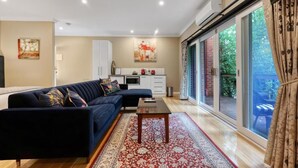 Una sala de estar moderna con un sofá azul oscuro, una mesa de centro de madera y una alfombra roja con estampado. Se ve un área de cocina con armarios blancos y una mesa de comedor a través de una puerta de vidrio.