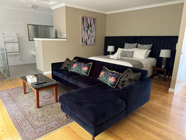 A modern hotel room with a dark blue sofa, a large bed with multiple pillows, a bedside table with a lamp, and a large mirror above the sink. - Wrenfield Cottages (Marysville)