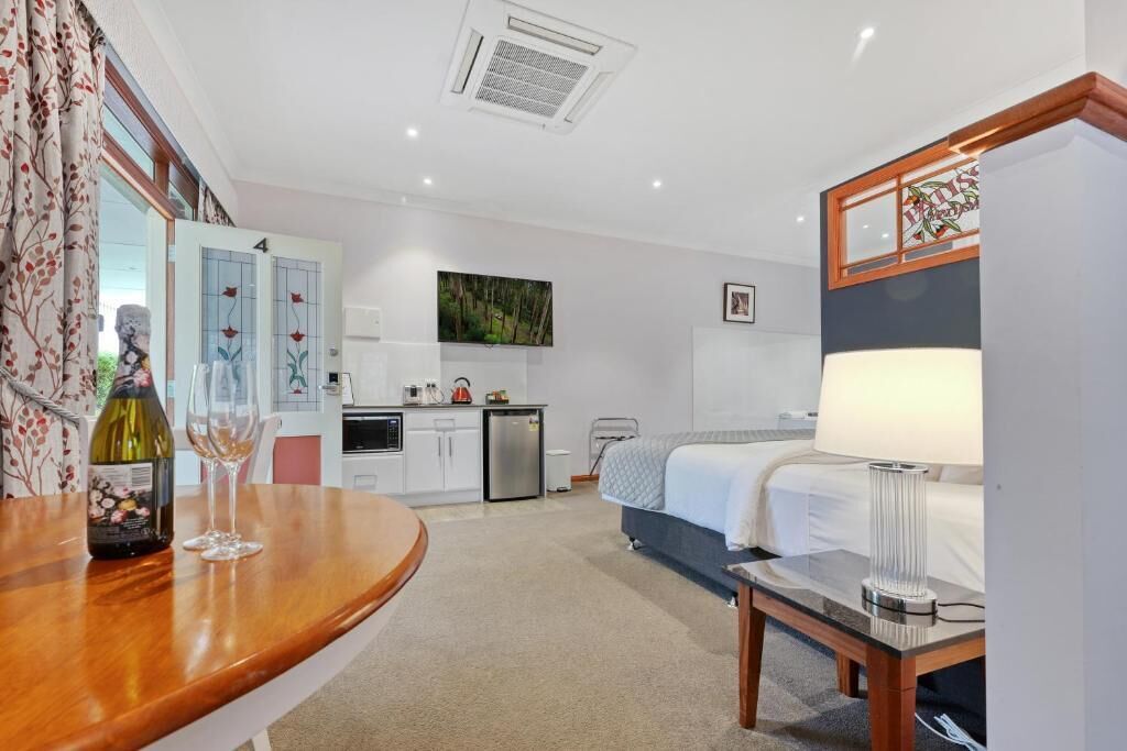 Quarto de hotel com cama, mesa de cabeceira, luminária e vista para a cozinha.