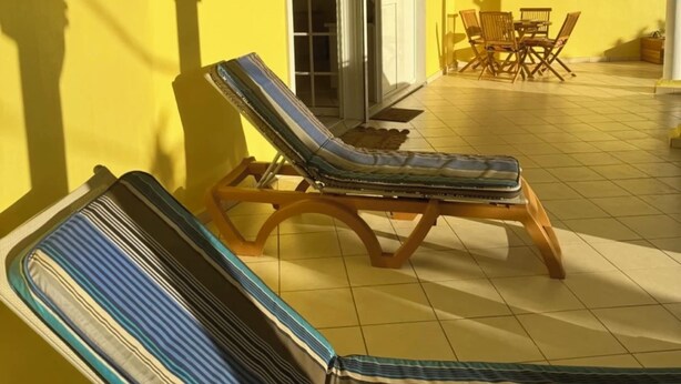 Eine sonnige Terrasse mit zwei gestreiften Liegestühlen und einem Tisch, gelben Wänden und einer Schiebetür.