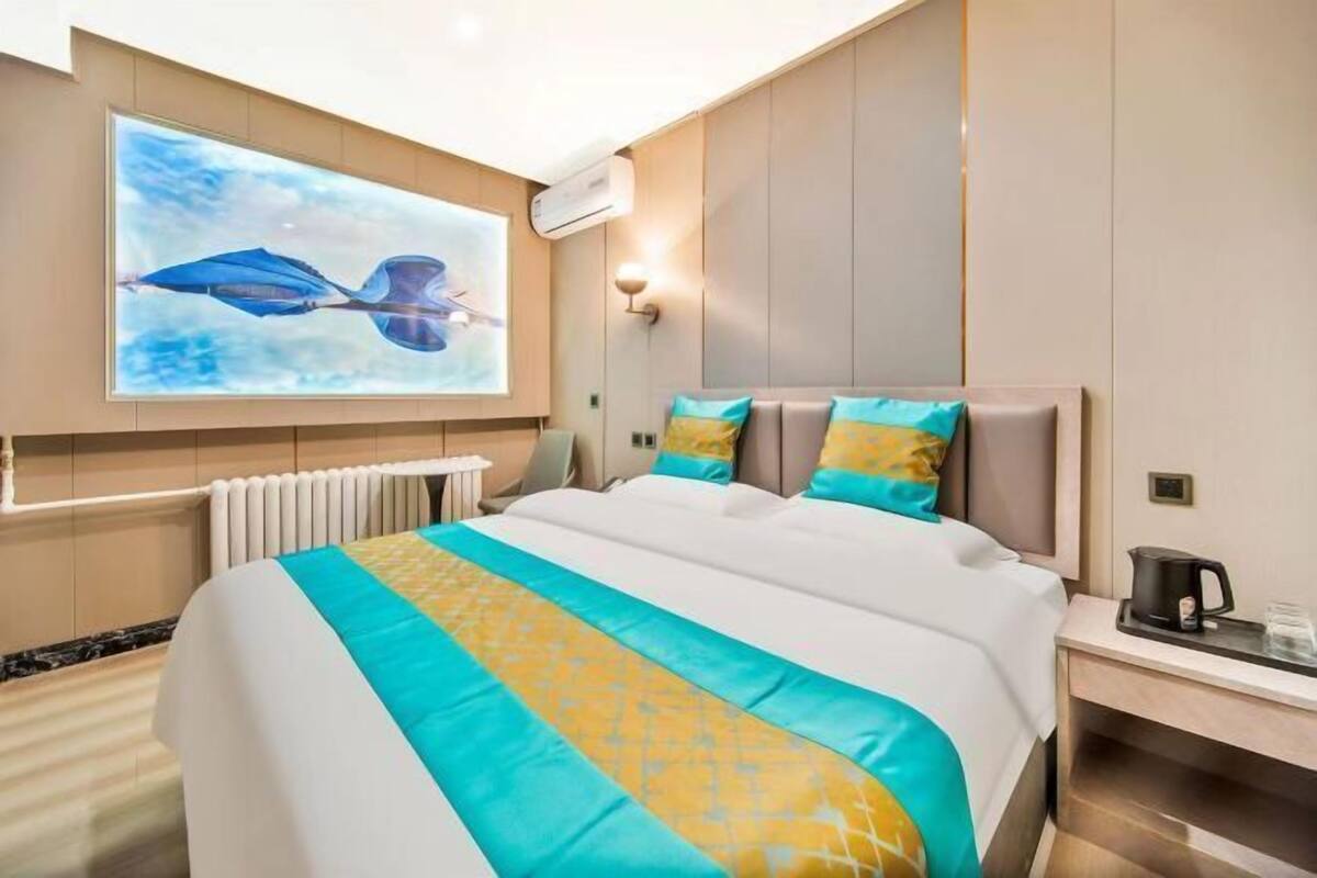 A hotel room with a large bed, a wall-mounted projector displaying a blue and white image, a bedside table with a coffee pot, and a radiator under the bed.