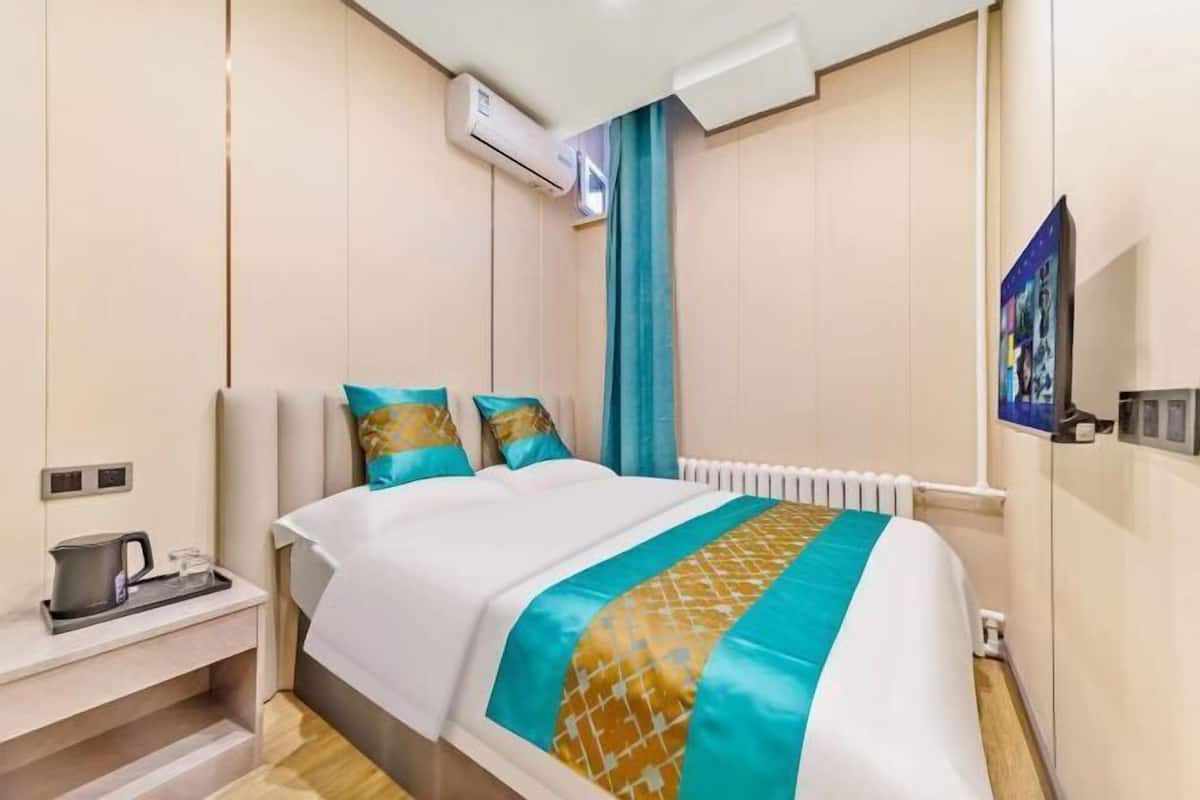 A hotel room with a bed, a nightstand with a kettle, a radiator, and a wall-mounted air conditioning unit.