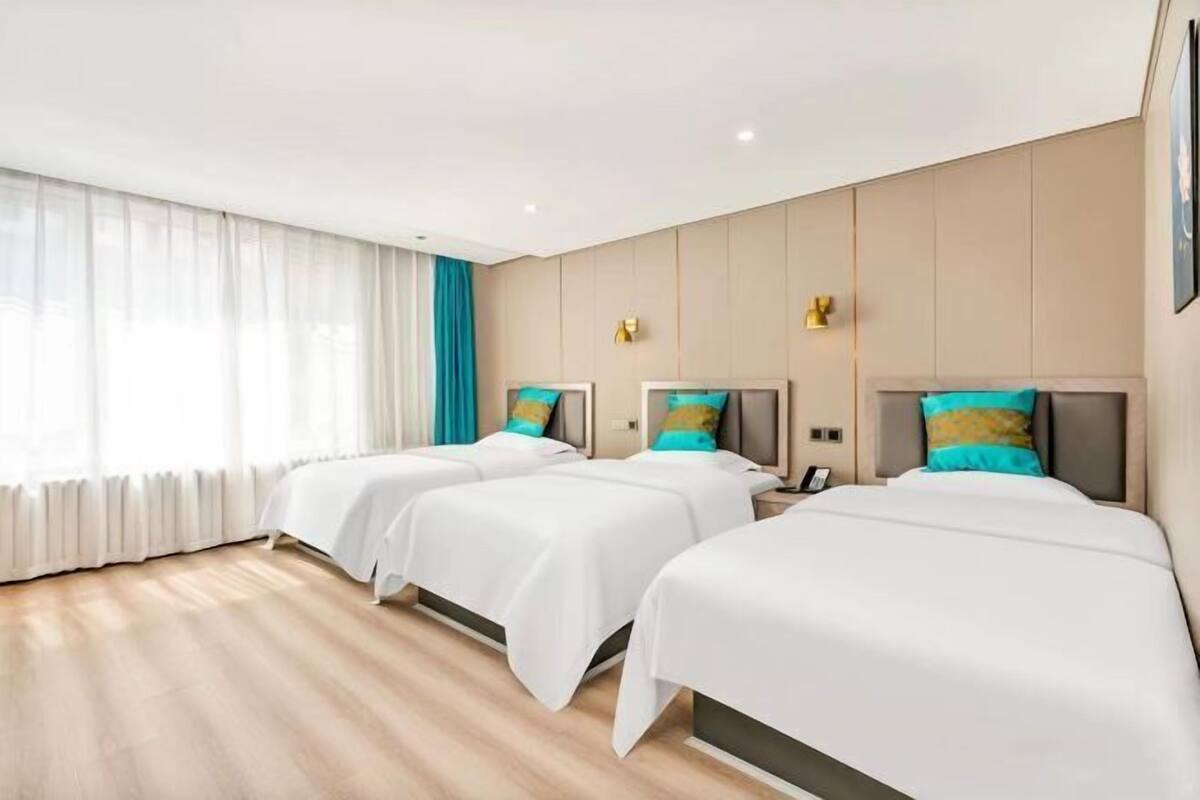 A hotel room with three beds, a large window with sheer curtains, and a light-colored wooden floor.