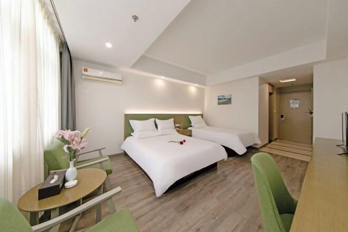 A hotel room with two beds, a small table with a vase of flowers, a green chair, and a wooden desk.