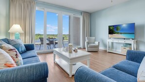 A living room with a sofa, a coffee table, a TV, and a balcony with a view of the beach.