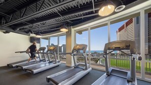 A gym with treadmills and a person using one, large windows with a view of the ocean and buildings, and a high ceiling with visible ductwork.