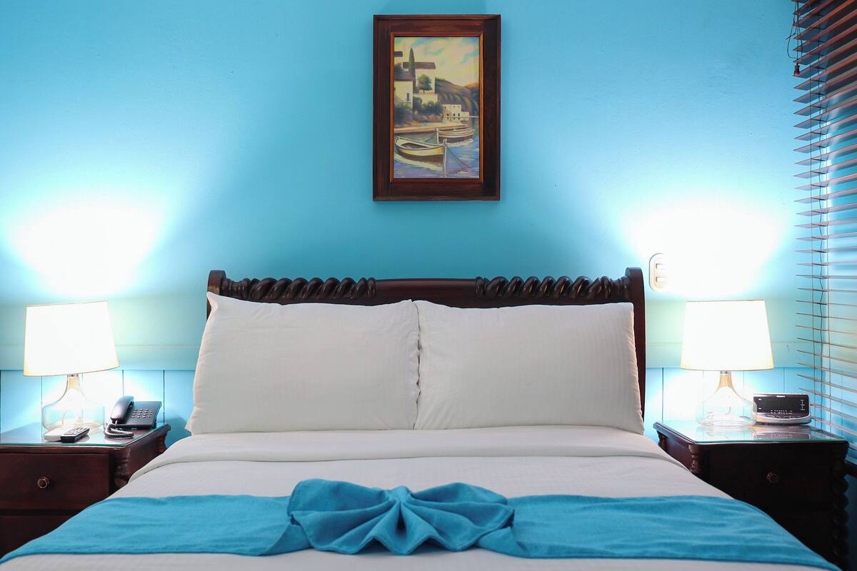Une chambre d’hôtel avec un lit, deux tables de chevet avec des lampes, un tableau au mur et une fenêtre avec des stores.