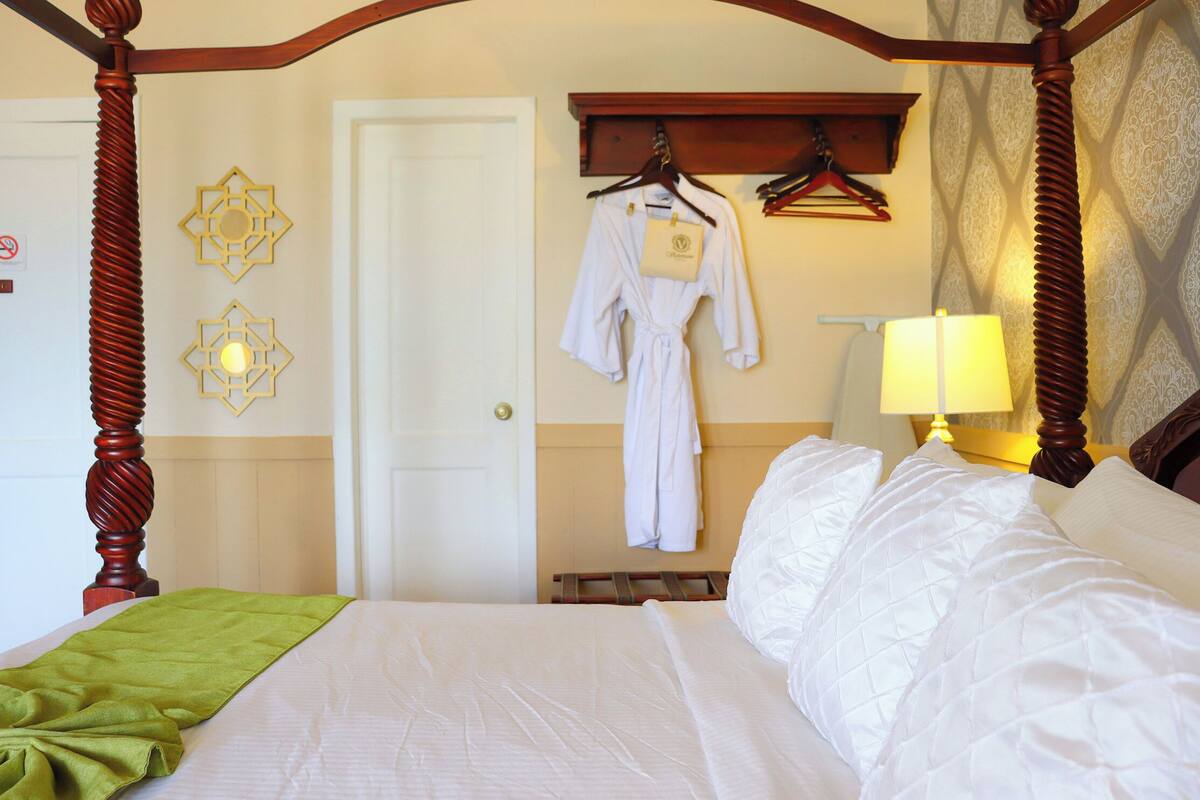 Une chambre d’hôtel avec un lit à baldaquin, un plaid vert, une robe accrochée au mur et une lampe de chevet.